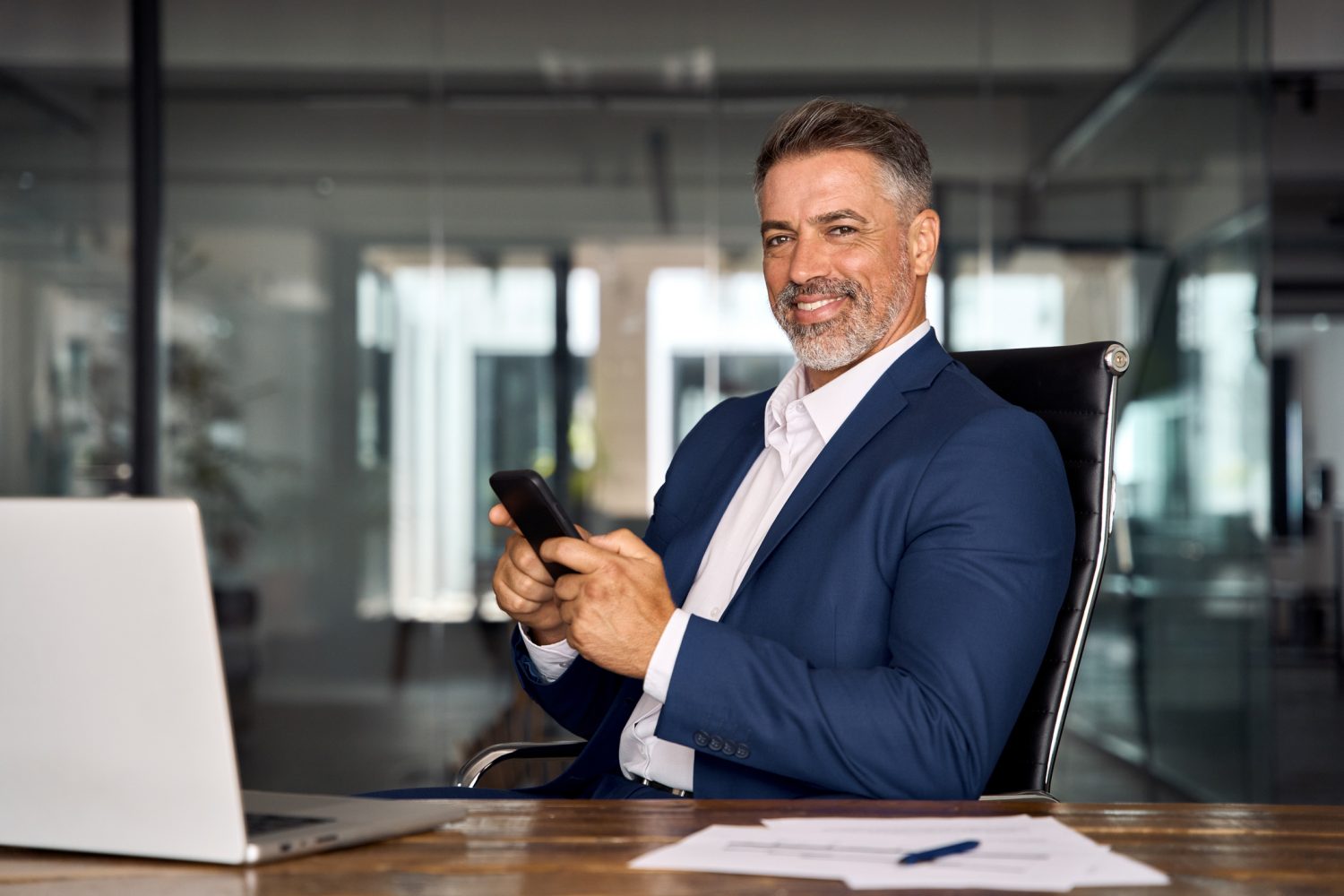 Middle aged Hispanic business manager ceo using cell phone mobile app, looking at camera. Smiling Latin or Indian mature man businessman holding smartphone sit in office working online in office.